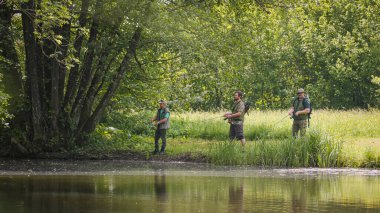 Nehir kıyısının yemyeşil doğasıyla çevrili üç balıkçı balık tutmayı seviyor. Açık hava hobisi ve aktivite konsepti.