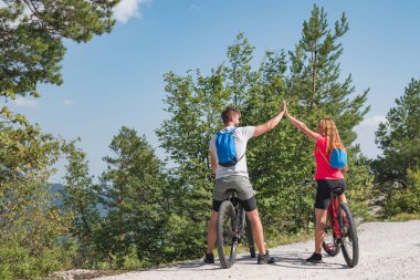 Genç çift muhteşem manzaranın tadını çıkarıyor, elektrikli dağ bisikletlerinin üzerinde oturmuş gülümsüyor ve birbirlerine beşlik çakıyorlar..