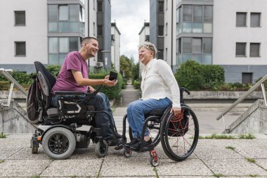 İki arkadaş, tekerlekli sandalye kullanıcıları sokakta gülümsüyor ve akıllı telefona bakıyor. Özürlü ve sosyal refah kavramları olan insanlar..
