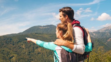 Bir çift genç dağcı, bir adam ve bir kadın, dağların manzarasına sarılıp, uzaklığı işaret ediyorlar.