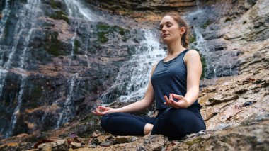 Sakin bir kız yoga yapıyor, sukhasana pozunda oturuyor arka planda güzel bir şelale mırıldanırken. Huzur, doğa ve sağlıklı yaşam tarzları..