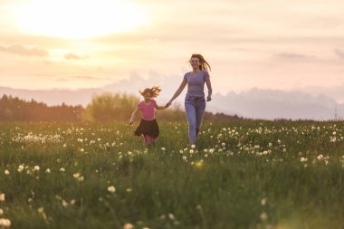 Mutlu ve gülümseyen anne ve küçük kızı güzel batan güneşin altında bir çayırda el ele koşuyorlar..