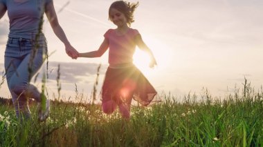 Mother and her little daughter running holding hands across the beautiful green grassland at sunset. clipart