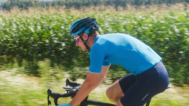 Beyaz bir yol bisikletçisinin yan görüntüsü, mavi spor giyimli, kasklı ve güneş gözlüklü, yarış bisikleti süren