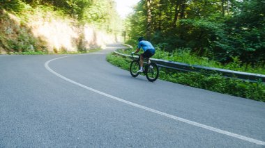 Male road racing cyclist in a blue sports jersey, with helmet and glasses, sprinting up a hill through the forest.