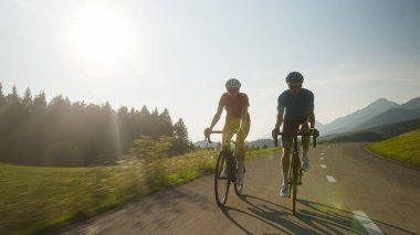 Bir kadın ve bir erkeğin ön görüntüsü, spor giyimli profesyonel yarış bisikletçileri, asfalt bir yolda güzel yeşil doğada ilerliyorlar..