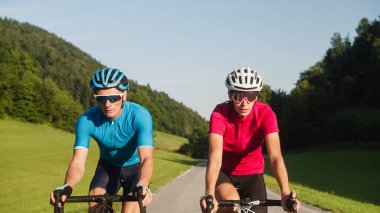 Bir kadın ve bir erkeğin ön görüntüsü, spor giyimli profesyonel yarış bisikletçileri, asfalt bir yolda güzel yeşil doğada ilerliyorlar..