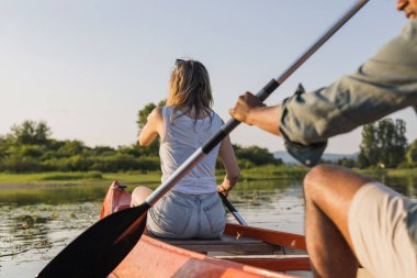 Gün batımında sakin göl suyunda kano süren çift, çevreleyen yeşil doğa ve renkli gökyüzü manzarasının sessizliğinin tadını çıkarıyor..