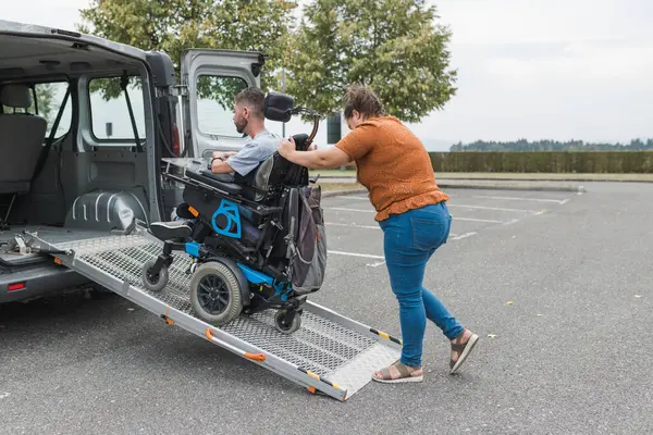 stock image Female assistant helping a man with disability to enter an accessible vehicle, pushing him over a wheelchair car ramp.