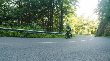 Male road racing cyclist in a blue sports jersey, with helmet and glasses, sprinting up a hill through the forest.