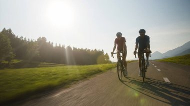 Front view of two people, a male, and female racing cyclists ride on a paved road through beautiful mountain nature at sunset