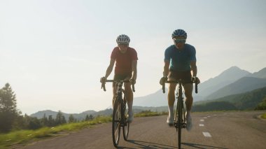 Bir kadın ve bir erkeğin ön görüntüsü, spor giyimli profesyonel yarış bisikletçileri, asfalt bir yolda güzel yeşil doğada ilerliyorlar..