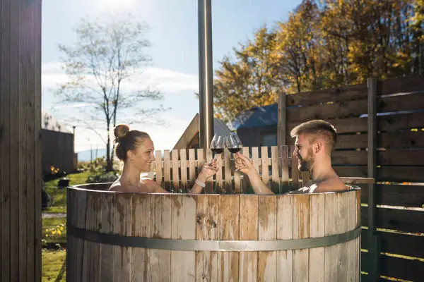 Genç bir çift bornozlarını çıkarıyor ve özel bir açık hava odunuyla yanan jakuziye giriyor. Glamping ve lüks kamp konsepti.