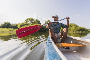 Genç adam kano yapıyor, kürek çekiyor ve göl suyunda kano gezdiriyor, doğal çevrenin tadını çıkarıyor..