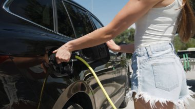 Sıradan bir yaz kıyafeti giymiş bir kız, bir soket açıyor ve şehir caddesindeki bir EV şarj istasyonunda siyah bir elektrikli arabaya şarj aleti takıyor.