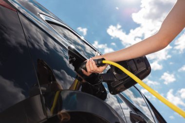 Mahalledeki bir elektrik istasyonunda zarif siyah EV arabasını şarj eden genç bir kadın. Temiz ve alternatif enerji kavramı.
