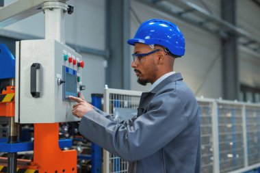 Afrika kökenli Amerikalı mühendis, gri bir iş ceketi ve mavi bir kask giyiyor, fabrikada bir makineyi kontrol ediyor ve programlıyor..