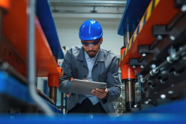 Mavi kasklı Afro-Amerikan mühendis elinde tablet, fabrikada makine teftişi yapıyor, verimliliği ve doğruluğu kontrol ediyor..