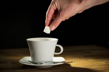 Erkek el, şeker küplerini beyaz bir kahve bardağına koyuyor ve çay kaşığıyla içkiyi karıştırıyor, yakın çekim. Sabah keyif konsepti.