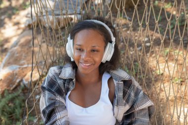 Kaygısız genç Afrikalı Amerikalı kadın hamakta dinleniyor, kablosuz kulaklıkla müzik dinlemekten ve etrafındaki güzel doğadan keyif alıyor..