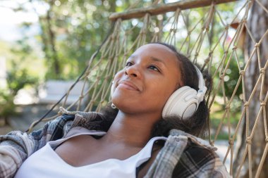 Afro-Amerikalı bir kadın hamakta dinleniyor, akıllı telefondan müzik seçiyor ve kulaklıkla favori şarkıları dinliyor..