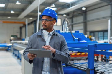Afrika kökenli Amerikalı mühendis elinde bir tabletle üretim hattında yürüyor ve otomatikleştirilmiş endüstriyel süreçleri izliyor..