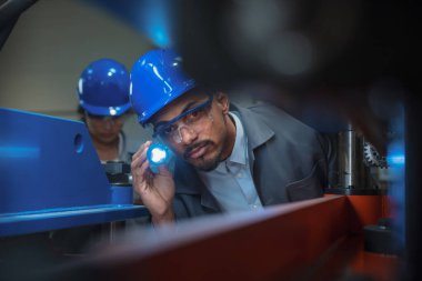 Makine mühendisi, Afrikalı Amerikalı adam el fenerini içerideki makine kontrolü için kullanıyor, Hintli bayan meslektaşıyla birlikte çalışıyor..