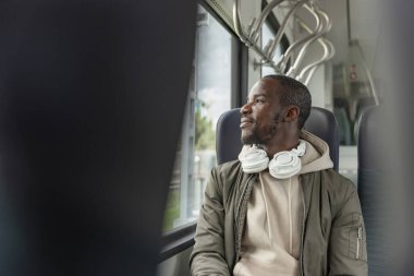 Gülümseyen Afrikalı Amerikalı adam rahat ve rahat tren yolculuğundan zevk alıyor, bir pencerenin kenarında oturuyor ve kulaklıkla müzik dinliyor..