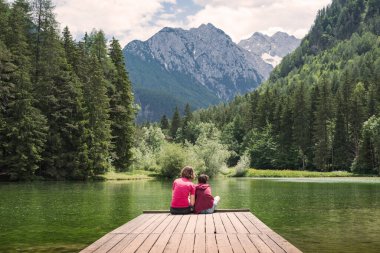 Anne rıhtımda oturmuş oğluna sarılıyor, görkemli dağ ve göl manzarasının tadını çıkarıyor. Aile zamanı ve tatil kavramları.