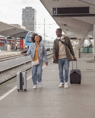 Tren istasyonunda yürüyen ve bavullarını taşıyan Afro-Amerikalı çift tren yolculukları için hazırlar. Gezgin yaşam tarzı konsepti.