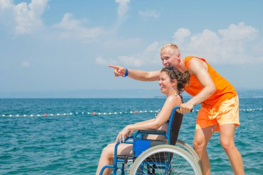 Engelli gülümseyen bir kadın ve asistanı tekerlekli sandalyesinin hareket etmesine yardım ediyor, sahilde ve deniz ufkunda eğleniyor..