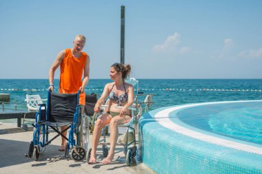 Deniz oteli cankurtaranı engelli bir kadına yardım ediyor erişilebilir hizmetler ve tesisler, havuz sandalyesi kaldırma, ve tekerlekli sandalye, tatil köyünde..