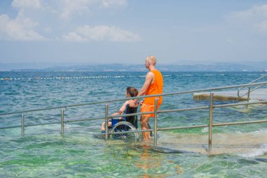 Erkek asistan, engelli bir kadını, yaz tatilinde dinlenmek için, dalgalı deniz kenarındaki ulaşılabilir sahil rampasına itiyor..