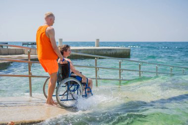 Engelli bir kadın, deniz suyuna bir cankurtaran yardımcısının yardımıyla erişilebilir tekerlekli sandalye plaj rampasından giriyor..