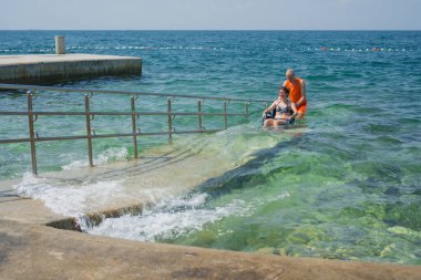 Engelli bir kadın, asistanının yardımıyla deniz suyundan çıkıyor, erişilebilir tekerlekli sandalye plaj rampasından geçiyor..