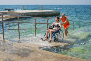 Rahatlamış, engelli bir kadın, bir asistanın yardımıyla deniz suyundan çıkıyor, erişilebilir tekerlekli sandalye plaj rampasından geçiyor..