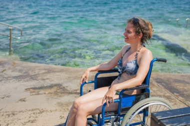 Bikinili gülümseyen bir kadın, deniz kenarında mavi tekerlekli sandalyesinde oturuyor, yaz tatilinde, bir deniz banyosundan sonra güneşin tadını çıkarıyor..