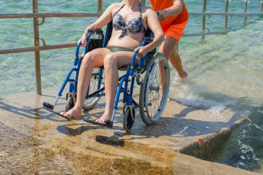 Engelli gülümseyen bir kadın ve asistanı, tekerlekli sandalyesinin hareket etmesine yardım eden bir plaj görevlisi, güneşli bir yaz gününde güzel bir deniz manzarasının keyfini çıkarıyorlar..