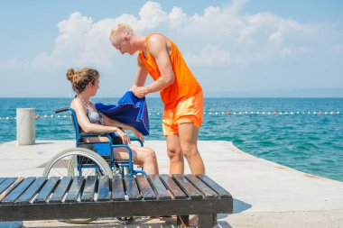 Plaj cankurtaranı engelli birine yardım ediyor. Deniz kenarında güneşli bir günde tekerlekli sandalyede oturan genç bir kadın için mavi havlu tutuyor..