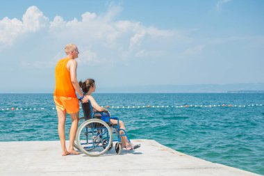 Birlikte, engelli gülümseyen bir kadın, tekerlekli sandalye kullanıcısı ve sahil görevlisi asistanı yaz güneşinin altındaki güzel deniz manzarasının tadını çıkarıyorlar..