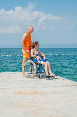 Birlikte, engelli gülümseyen bir kadın, tekerlekli sandalye kullanıcısı ve sahil görevlisi asistanı yaz güneşinin altındaki güzel deniz manzarasının tadını çıkarıyorlar..