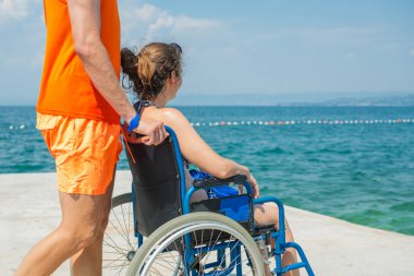 Birlikte, engelli gülümseyen bir kadın, tekerlekli sandalye kullanıcısı ve sahil görevlisi asistanı yaz güneşinin altındaki güzel deniz manzarasının tadını çıkarıyorlar..