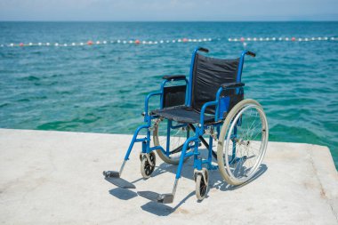 Engelliler için boş mavi çelikli tekerlekli sandalye, deniz kenarına yerleştirilir, güzel sahil çevresine erişim sağlanır..