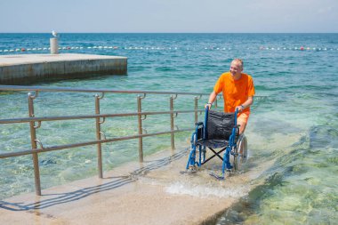 Gülümseyen cankurtaran, denizden gelen boş bir tekerlekli sandalyeyi sahil rampası boyunca itiyor, ulaşılabilir bir turizm hizmeti, bir çevre, ve bir yaz durağı, ön manzara..