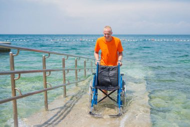 Gülümseyen cankurtaran, denizden gelen boş bir tekerlekli sandalyeyi sahil rampası boyunca itiyor, ulaşılabilir bir turizm hizmeti, bir çevre, ve bir yaz durağı, ön manzara..