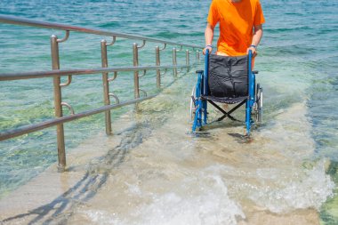 Sahil rampası boyunca denizden gelen boş bir tekerlekli sandalyeyi iten cankurtaran, ulaşılabilir bir turizm hizmeti, bir çevre ve bir yaz durağı, ön manzara..