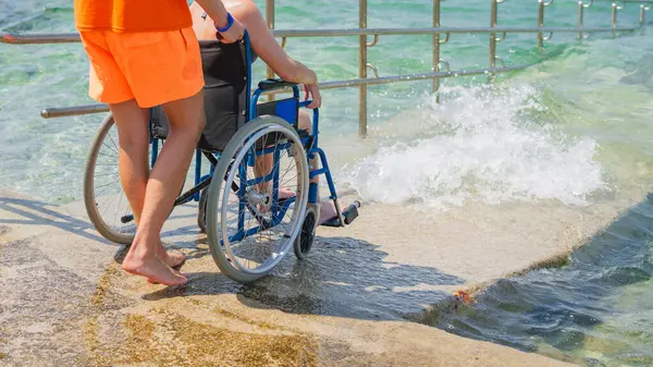 Engelli bir kadın, deniz suyuna bir cankurtaran yardımcısının yardımıyla erişilebilir tekerlekli sandalye plaj rampasından giriyor..