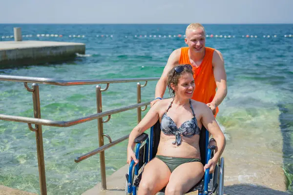 Rahatlamış, engelli bir kadın, bir asistanın yardımıyla deniz suyundan çıkıyor, erişilebilir tekerlekli sandalye plaj rampasından geçiyor..