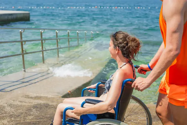 Yardımsever bir erkek asistan, mavi denize doğru uzanan ulaşılabilir sahil rampası boyunca, tekerlekli sandalyeli bir kadını ittiriyor..