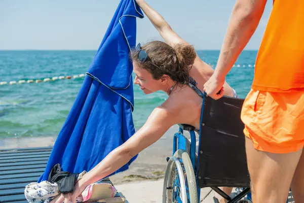 Plaj cankurtaranı engelli birine yardım ediyor. Deniz kenarında güneşli bir günde tekerlekli sandalyede oturan genç bir kadın için mavi havlu tutuyor..